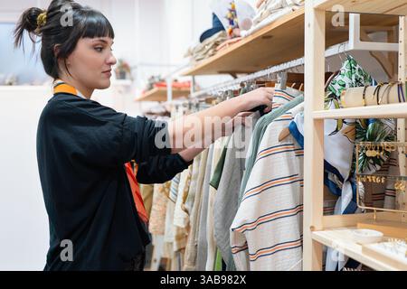 Una donna sfoglia abiti colorati in una boutique moderna, circondata da abbigliamento e accessori ordinati su scaffali in legno, che riflettono uno stile elegante Foto Stock