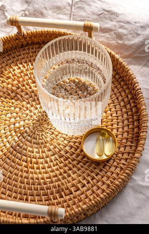 Dall'alto un bicchiere trasparente di acqua e integratori, tra cui capsule Omega 3 e vitamina D3 poste su un vassoio di rattan, che incarna una conce olistica per la salute Foto Stock