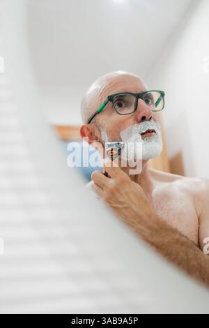 Un uomo maturo si vede radersi in un bagno ben illuminato, usando un rasoio mentre si guarda allo specchio. L'ambiente trasmette un senso di cura personale e quotidiano Foto Stock