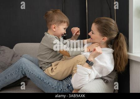 Una madre e un figlio gioiosi condividono un momento di gioco sul divano. Il bambino siede sulla mamma in grembo, sorridente e godendo del tempo di qualità in una vita accogliente Foto Stock