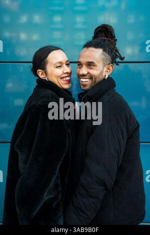 Una felice coppia multiculturale vestita con eleganti cappotti invernali gode di un momento felice insieme, su uno sfondo blu moderno, che mette in risalto l'amore e l'amore Foto Stock