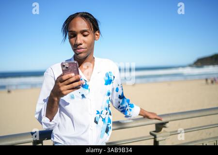 Un giovane uomo, con una camicia blu e bianca vivace, controlla il suo smartphone mentre si appoggia su una ringhiera da spiaggia. La scena riflette orgoglio e connettività Foto Stock