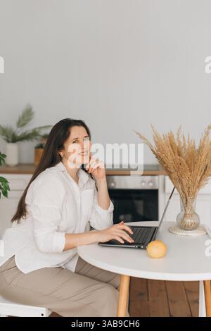 Un nutrizionista siede in una cucina moderna, sorridendo mentre lavora su un notebook. Un limone e una pianta decorativa sono sul tavolo, creando un voi professionale Foto Stock