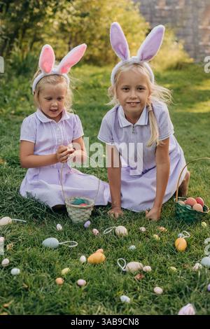 Due bambini che indossano orecchie da coniglio partecipano a una gioiosa caccia all'uovo di Pasqua in una giornata di sole. Raccolgono uova colorate in cesti, circondati dal verde A. Foto Stock
