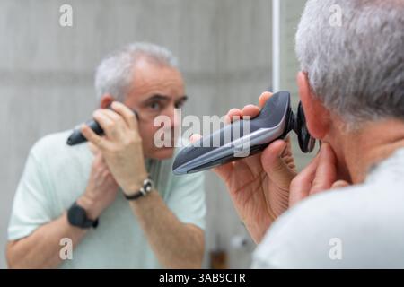 Un uomo maturo con i capelli grigi si rasa con cura il viso e le basette utilizzando un rasoio elettrico mentre si guarda allo specchio. La sua espressione focalizzata Highli Foto Stock