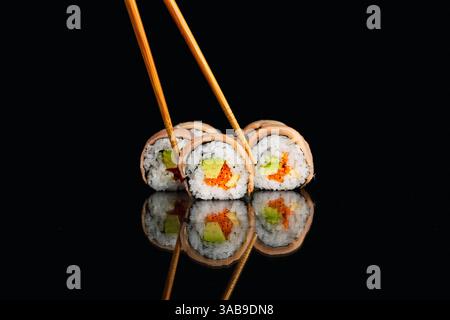 I panini di sushi con avocado, carote e altri ingredienti vivaci sono elegantemente preparati con bacchette. Poggiano su una superficie nera lucida, creando Foto Stock