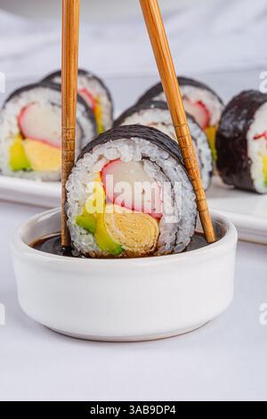 Primo piano di un sushi roll tenuto da bacchette, immerso in una ciotola di salsa di soia su sfondo bianco. Con ingredienti colorati come il bastoncino di granchio, l'avoca Foto Stock