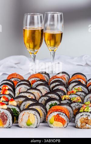 Un elegante assortimento di sushi roll con condimenti vivaci abbinati a due bicchieri di spumante, su sfondo bianco. Perfetto per una celebra Foto Stock
