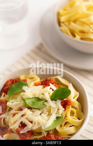 Un primo piano di spaghetti conditi con salsa di pomodoro, formaggio grattugiato e spinaci freschi in una ciotola. Lo sfondo presenta una ciotola di pasta semplice. Ideale Foto Stock