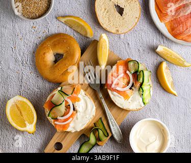 Vista dall'alto di un bagel di sesamo ricoperto da formaggio cremoso, salmone affumicato, fette di cetriolo e cipolla, circondato da cunei di limone su un rustico legno Foto Stock