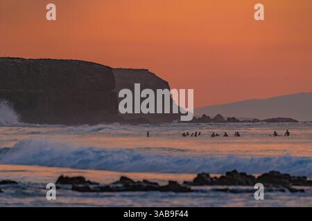 I surfisti si godono le onde al tramonto a Fuerteventura, Isole Canarie. Il cielo arancione e le aspre scogliere creano uno sfondo perfetto per questa vivace stagione Foto Stock