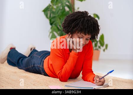 Donna afroamericana sdraiata sul pavimento che scrive su un taccuino con penna a casa Foto Stock