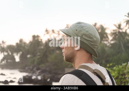 Uomo in cappello e zaino che guarda la tranquilla costa tropicale durante l'alba, guardando lontano. Palme e rocce sullo sfondo. Vacanze estive di viaggio Foto Stock