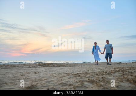 Una coppia cammina mano nella mano lungo una spiaggia sabbiosa durante un tranquillo tramonto. I due appaiono gioiosi e rilassati, abbracciando la tranquilla atmosfera serale Foto Stock