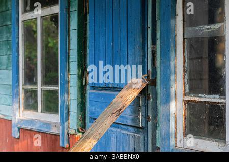 Una casa di legno intemprata caratterizzata da una porta blu brillante fissata con una tavola di legno e finestre d'epoca. Le texture rustiche e i colori vivaci evocano un sen Foto Stock