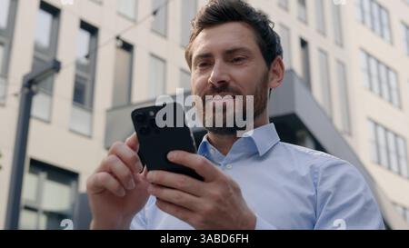 Sorridente positivo uomo d'affari caucasico di mezza età uomo d'affari ragazzo maschio datore di lavoro imprenditore lavoratore in città vicino all'edificio di uffici utilizzando il telefono cellulare Foto Stock