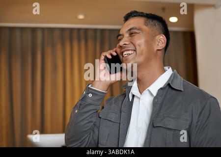 Uomo sorridente che parla al telefono mentre è in piedi nell'elegante hall dell'hotel Foto Stock