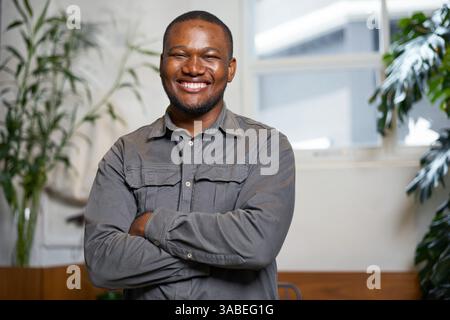 Uomo sicuro di sé sorridente con le braccia attraversato in un ambiente d'ufficio luminoso e moderno Foto Stock