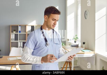 Ritratto di un giovane medico maschile in uniforme medica blu che lavora nel suo ufficio con blocco note. Foto Stock