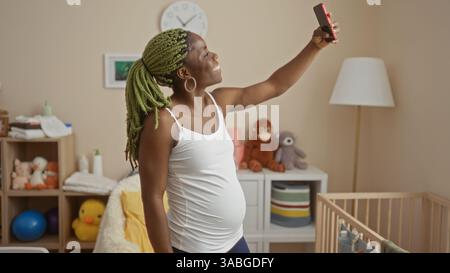 Donna con trecce che scatta selfie in un'accogliente camera da letto con culla che mostra momenti gioiosi di gravidanza e vita domestica Foto Stock