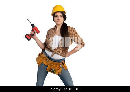 Una determinata lavoratrice edile che indossa un elmetto giallo e una cintura portautensili, reggendo con sicurezza un trapano mentre si trova contro una schiena bianca Foto Stock