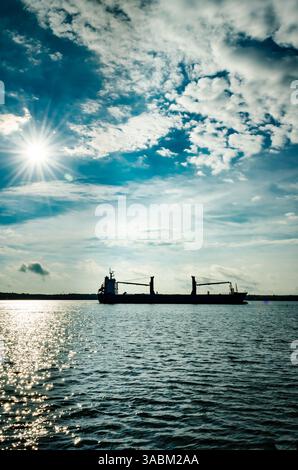 La nave da carico si staglia con un sole stellato dietro di essa contro un cielo blu nuvoloso e un'acqua blu scuro Foto Stock