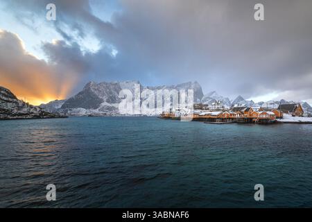 Sakrisoy auf den Lofoten in Norwegen unter dichter Wolkendecke mit Blick auf gelbe Rorbuer und goldenen Sonnenstrahlen, Die den Himmel durchbrechen Foto Stock