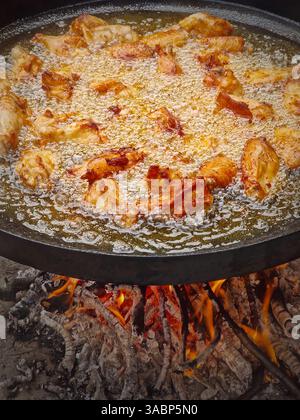 Ali di pollo che friggono all'aperto in una grande padella sul fuoco. Bollire l'olio vegetale crea bolle mentre cuocere la carne sotto la fiamma Foto Stock