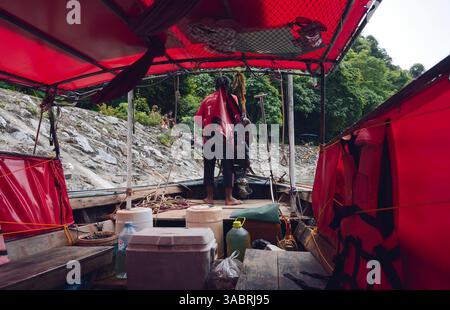 Il capitano della barca a coda lunga si prepara ad attraccare sotto i vivaci teloni rossi vicino a una costa rocciosa artificiale Foto Stock