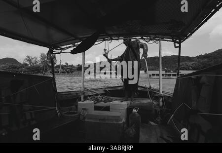 Il capitano della barca a coda lunga si trova al timone mentre la nave si avvicina a un molo in bianco e nero Foto Stock