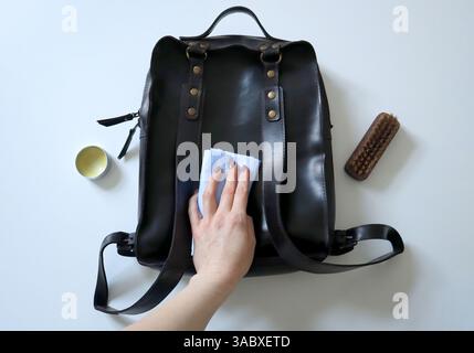 Zaino da donna in pelle di cavallo pazzo. Sta tenendo la spugna. Lo zaino è nero, sul retro. Hardware dorato. Cera e spazzolina per la pulizia nelle vicinanze. Sfondo bianco. Foto. Cura dei prodotti in pelle Foto Stock