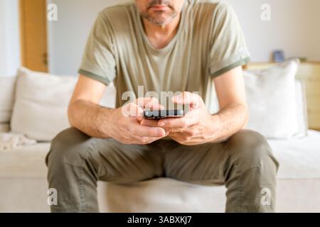 Uomo seduto sul divano con abiti casual che reggono lo smartphone. Messa a fuoco selettiva. Foto Stock