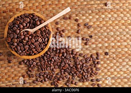 Prospettiva dall'alto verso il basso dei chicchi di caffè appena tostati in una ciotola di legno, con cucchiaio su un tappeto intrecciato testurizzato Foto Stock