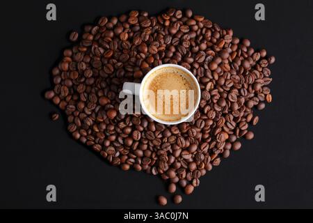 Vista dall'alto di una tazza bianca di espresso appena estratto circondato da chicchi di caffè tostati marroni su una superficie scura con spazio per copiare Foto Stock