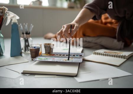 Le mani di un artista da primo piano mescolano l'acquerello per dipingere in un notebook in studio da pavimento con spruzzatore e tazze Foto Stock