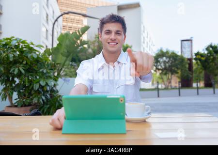 Sorridente uomo caucasico che punta alla fotocamera mentre usa un tablet in un bar all'aperto Foto Stock