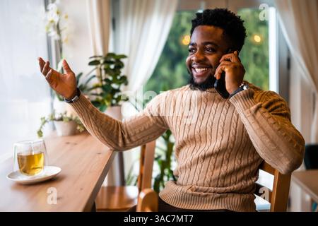 Un allegro uomo nero siede al bar, vestito in modo informale. Sta parlando al telefono cellulare. Foto Stock