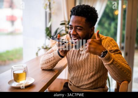Un giovane nero siede al bar, vestito in modo informale. Sta utilizzando lo smartphone e registrando un messaggio vocale. Foto Stock