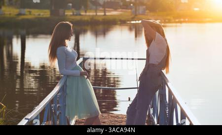 Le giovani donne indossano un abbigliamento casual su un molo di legno, crogiolandosi alla luce dorata del sole che si riflette sulle calme acque del lago Foto Stock