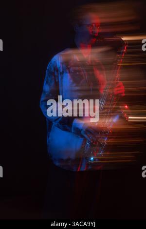 Ripresa dinamica verticale di un musicista maschile che indossa una camicia rilassata e si diverte a suonare il sassofono durante l'esibizione notturna in jazz club sotto luci al neon blu e rosse, effetto slow shutter Foto Stock
