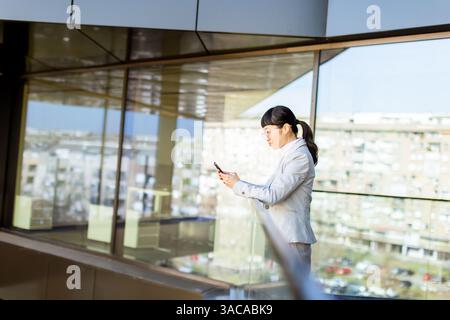 Una donna d'affari giapponese, vestita con un abito sartoriale, si stende su un balcone, utilizzando il suo smartphone mentre si gode una vista panoramica della città Foto Stock