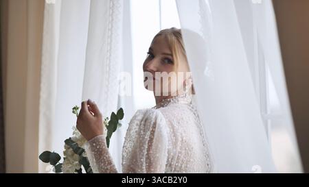Elegante sposa bionda con abito bianco, bouquet floreale, che guarda delicatamente sulle spalle vicino alla finestra luminosa Foto Stock
