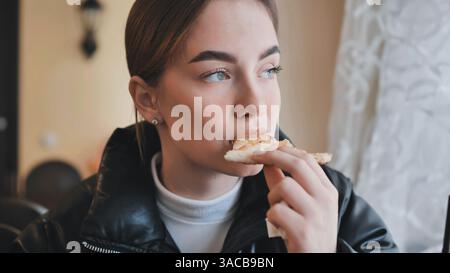 Una giovane donna che morde una fetta di pizza scadente, siede in un ristorante caldo, sorride mentre si indulge in un delizioso momento di pranzo Foto Stock