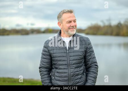 Un uomo maturo con una giacca imbottita scura si trova di fronte a un lago calmo, guardando lateralmente con un sorriso morbido e contento. Foto Stock