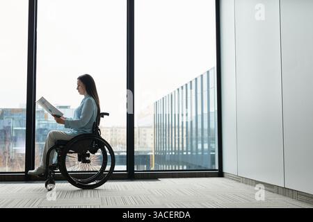 Ritratto grafico laterale di una giovane donna d'affari in sedia a rotelle che legge documenti da una finestra a tutta lunghezza in ufficio, spazio copie Foto Stock