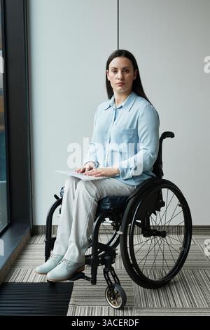 Ritratto verticale a tutta lunghezza di una giovane donna d'affari in sedia a rotelle che guarda la macchina fotografica mentre posa per una finestra in ufficio Foto Stock