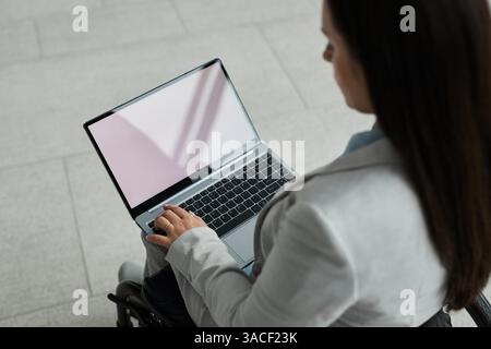 Ritratto ad angolo alto di una giovane donna d'affari in sedia a rotelle che utilizza un computer portatile con schermo bianco vuoto e spazio per copie Foto Stock