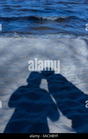 Un bel momento catturato: Ombre di due persone che condividono un bacio amorevole sulla spiaggia sabbiosa vicino all'acqua, dove le onde si tuffano dolcemente sulla costa Foto Stock