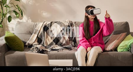 Ragazza con cappuccio rosa che utilizza il visore VR mentre assisti a un'esperienza coinvolgente in aula virtuale a casa. Futurismo, tecnologie moderne Foto Stock