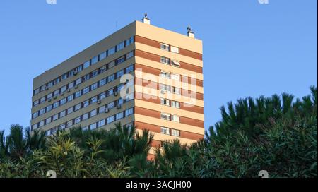 Moderno edificio residenziale a più piani e finestre, alto contro un cielo azzurro e circondato da lussureggianti alberi verdi Foto Stock